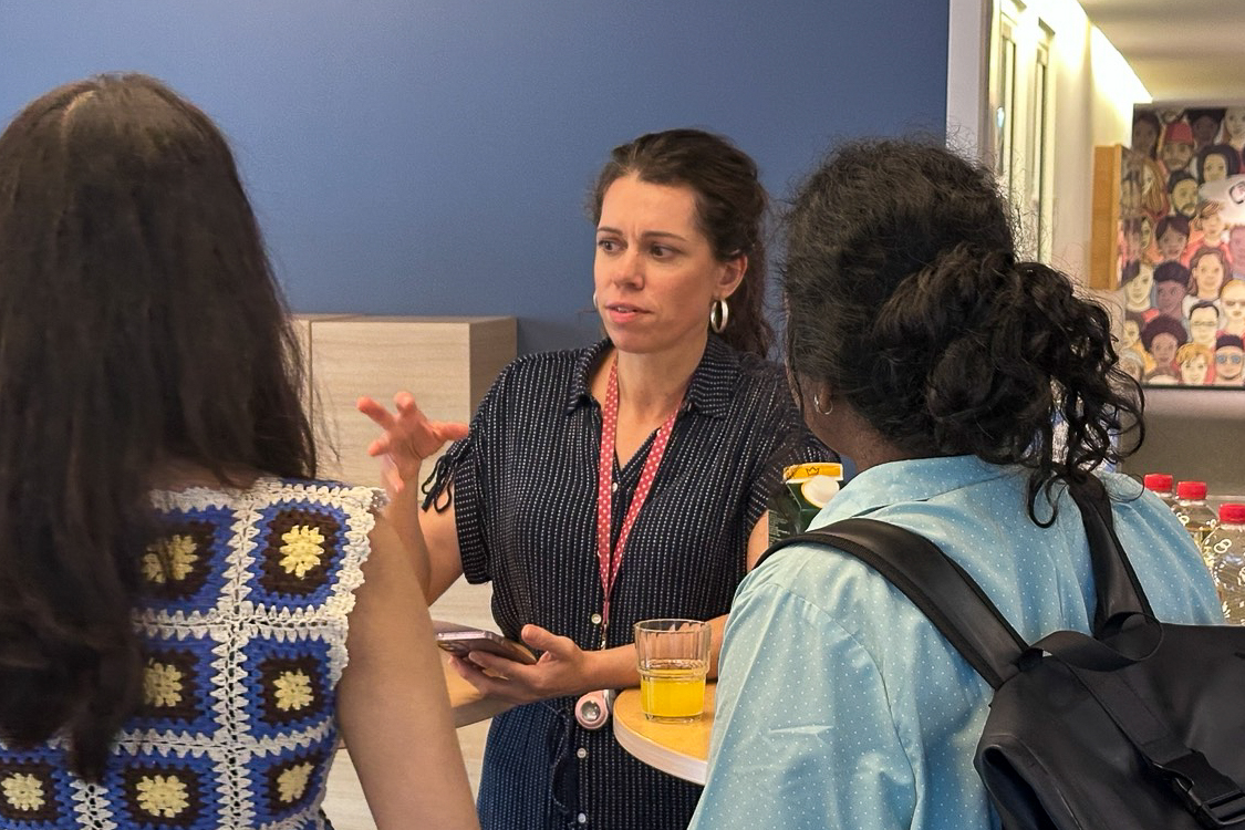 Woman with a lanyard speaking to two women, one wearing a crocheted top and the other with a backpack, near a table with drinks