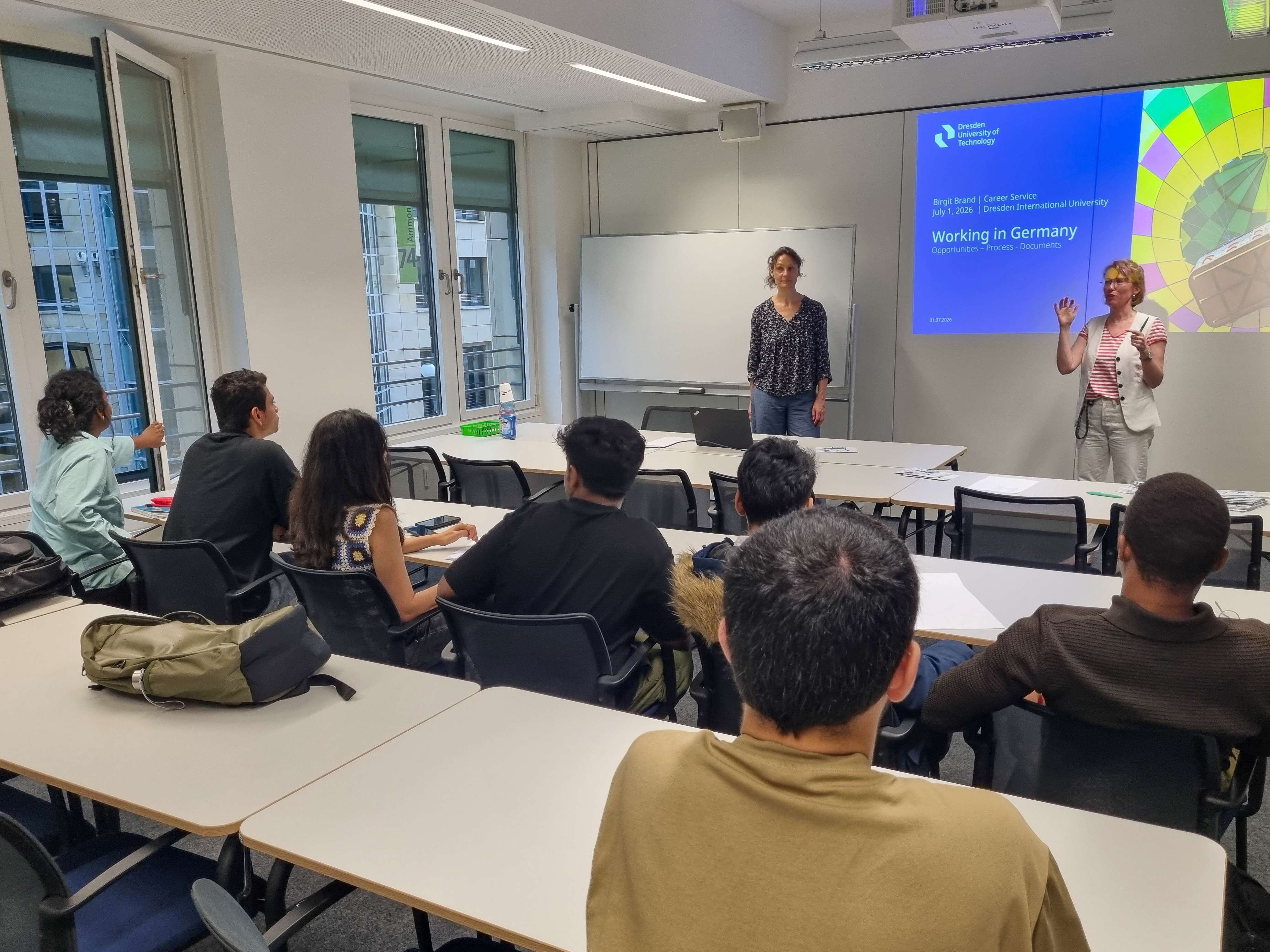 Classroom with students seated at tables facing two women presenting in front of a screen showing a slide titled 'Working in Germany'