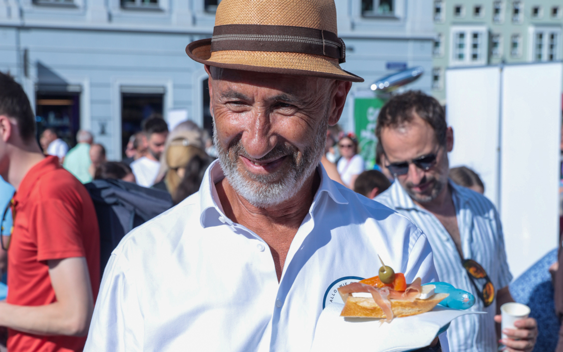 Älterer Mann mit Strohhut und weißem Hemd hält ein belegtes Brot mit Tomate und Oliven
