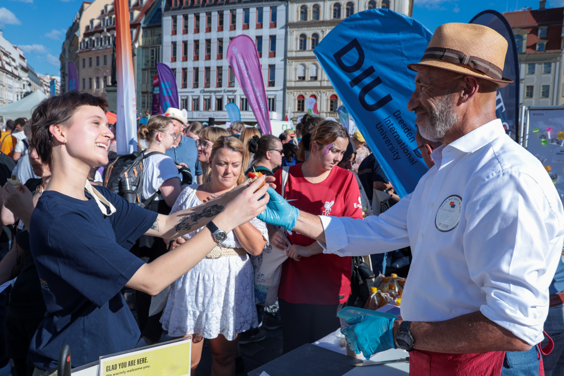 Älterer Mann mit Hut reicht einer jungen Frau mit kurzem Haar Essen am "Dresden is(s)t bunt" DIU Stand.