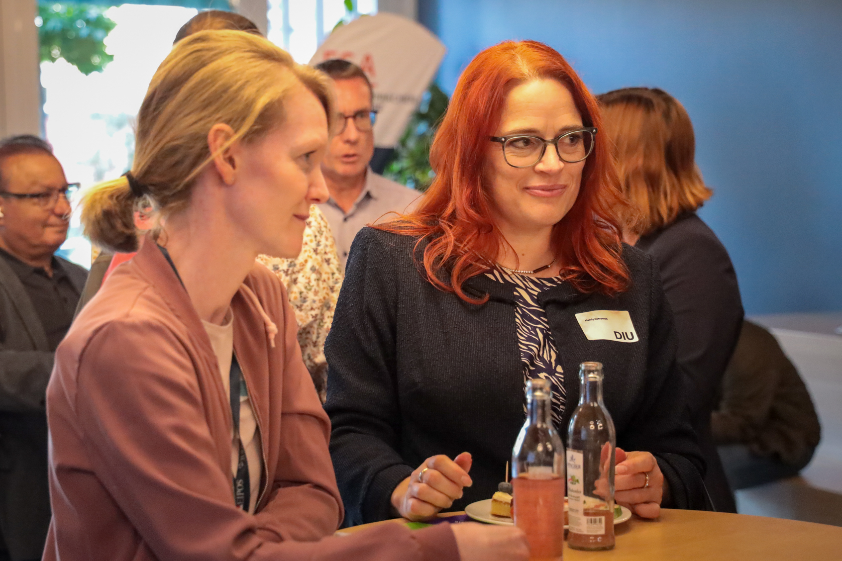 Zwei Frauen sitzen an einem Tisch, eine mit roten Haaren und Brille, die andere mit blondem Zopf, im Hintergrund mehrere Personen in einem Innenraum.