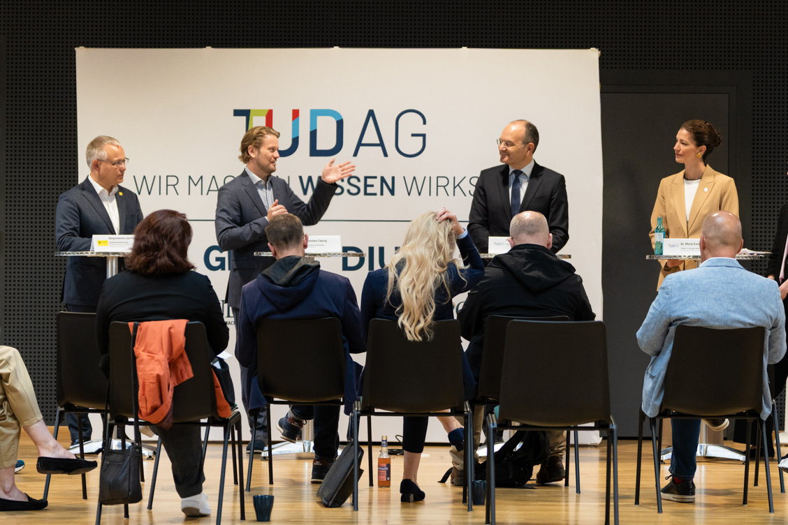 Vier Personen stehen an einem Podium vor einer Gruppe sitzender Zuhörer in einem hellen Raum mit einem Banner der TUDAG im Hintergrund