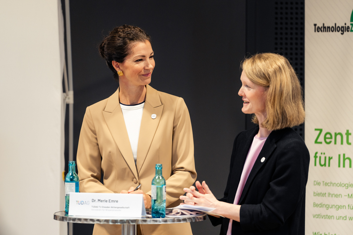 Zwei Frauen im Gespräch an einem Stehtisch mit Namensschild und Wasserflaschen, eine trägt beige Blazer, die andere schwarzen Blazer
