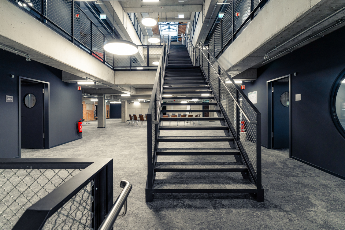 Industrieller Innenraum mit zentraler Metalltreppe, umlaufendem Geländer und mehreren Türen mit runden Fenstern in einem zweigeschossigen Gebäude