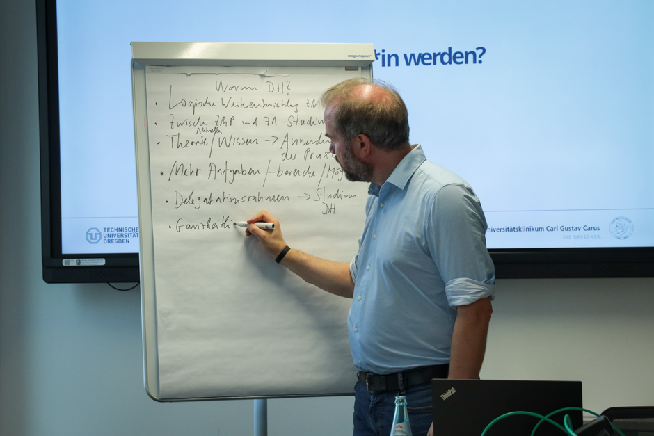 Mann mit Hemd steht vor einem Flipchart und schreibt darauf, im Hintergrund ein Banner mit Text und Logos.