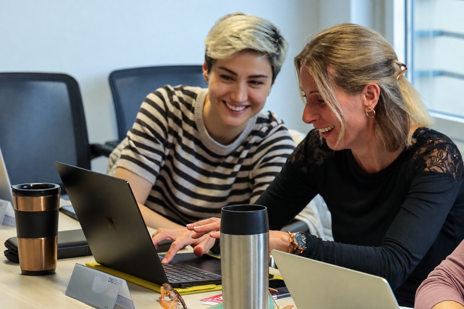 Zwei Frauen sitzen an einem Tisch und schauen lächelnd auf einen Laptop.