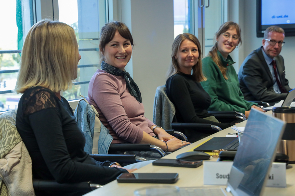 Fünf Personen sitzen an einem Konferenztisch, vier Frauen und ein Mann, sie blicken zueinander und lächeln.
