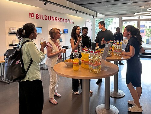 Group of six people standing around two round tables with bottles of juice and a bowl of fruit in a modern indoor space with 'Bildungscampus' sign on the wall