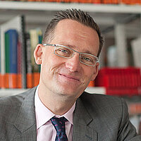 Mann mit Brille und Anzug sitzt lächelnd an einem Tisch mit aufgeschlagenen Büchern vor einem Bücherregal