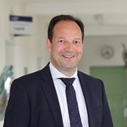 Portrait DIU Scientific Director Prof. Dr. R. Lasch: Man in a dark suit and striped tie standing in a corridor with windows and plants.
