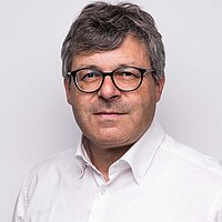 Mann mit grauen Haaren und Brille in weißem Hemd vor hellem Hintergrund
