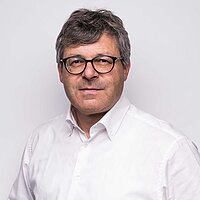 Mann mit grauen Haaren und Brille in weißem Hemd vor hellem Hintergrund