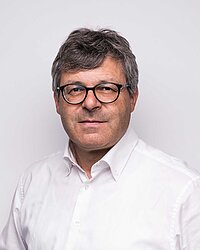 Mann mit grauen Haaren und Brille in weißem Hemd vor hellem Hintergrund