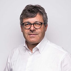 Mann mit grauen Haaren und Brille in weißem Hemd vor hellem Hintergrund