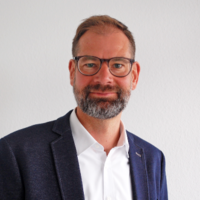 Mann in weißem Hemd und dunklem Sakko vor weißer Wand.