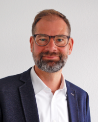 Mann in weißem Hemd und dunklem Sakko vor weißer Wand.