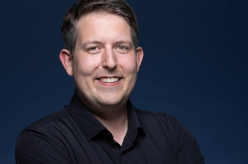 Smiling man with short dark hair wearing a black collared shirt against a dark blue background