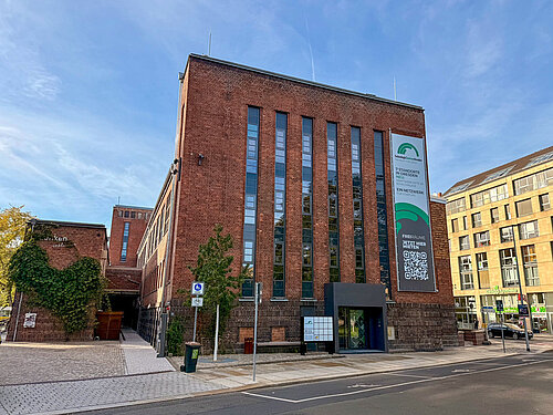 Mehrstöckiges rotes Backsteingebäude mit vertikalen Fenstern und großem Banner an der Fassade an einer Straße