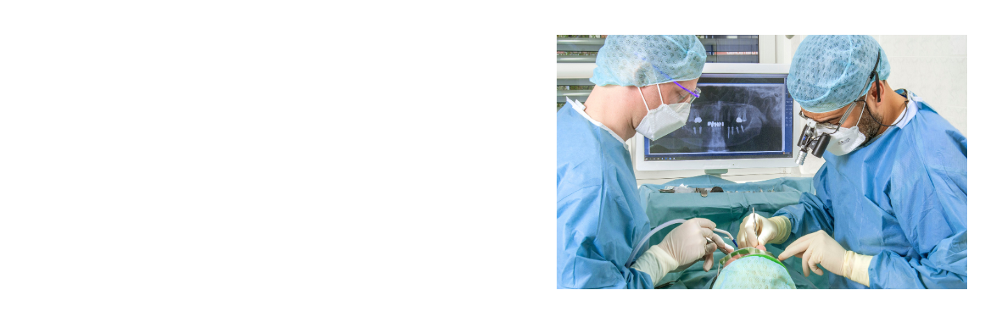 Two dental surgeons in full sterile protective gear perform a procedure on a patient, with a dental X-ray visible on a monitor in the background.