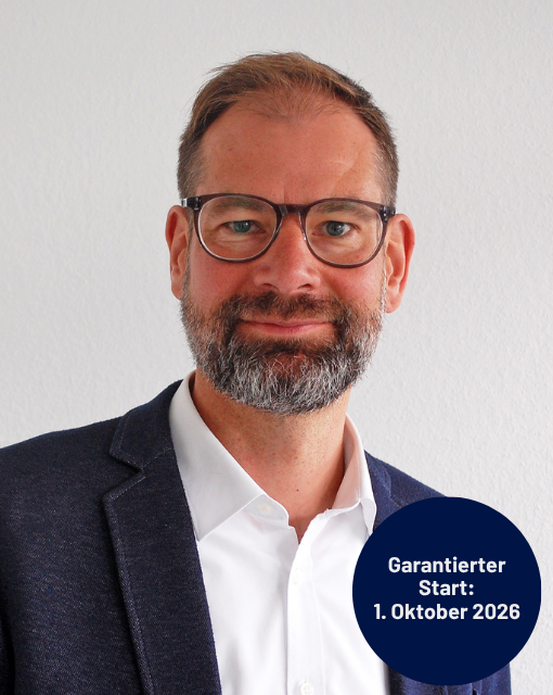 Mann mit Brille, Bart und dunklem Sakko vor weißer Wand, lächelnd