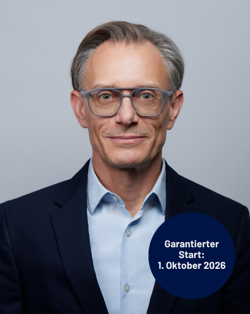Porträt eines älteren Mannes mit grauen Haaren, Brille und dunklem Sakko vor grauem Hintergrund mit Text in blauem Kreis 'Garantierter Start: 1. Oktober 2026'
