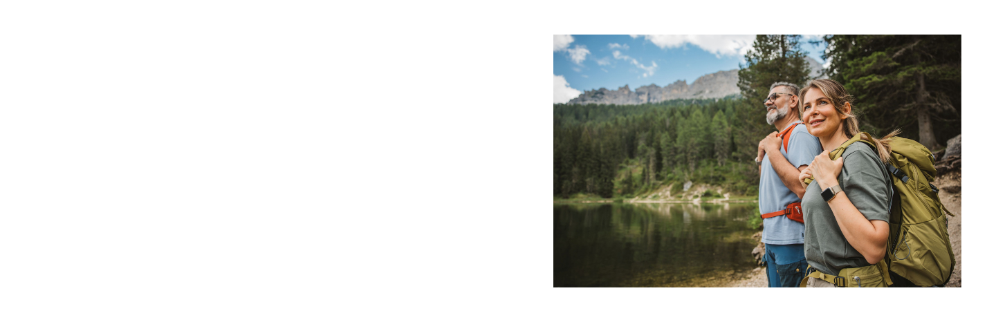 Zwei Wandernde mit Rucksäcken stehen an einem See vor bewaldeten Bergen unter blauem Himmel