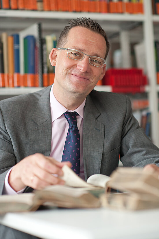 Mann mit Brille und Anzug sitzt lächelnd an einem Tisch mit aufgeschlagenen Büchern vor einem Bücherregal